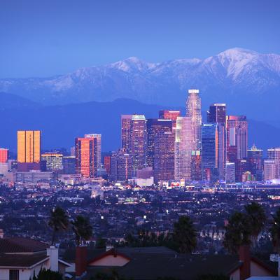 A view of downtown Los Angeles skyscrapers over snowy mountains during twilight.