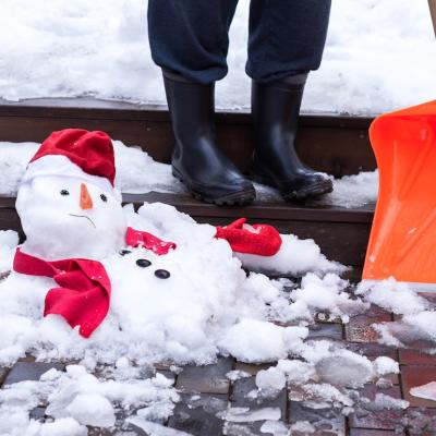 A snowman in red mittens, a scarf, and cap is melting outdoors on wet pavement next to a person's boots and shovel.