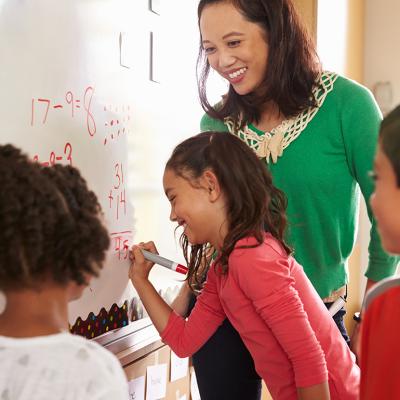 A grade school teacher happily looks over pupils answering a math exercise on the board.