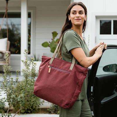 Travel nurse wearing olive green scrubs and carrying a tote bag looks over her shoulder with one hand on an open car door parked in driveway.