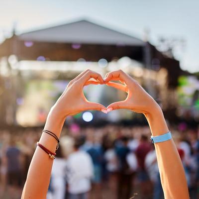 Concertgoer holds hands up to form a heart shape in the foreground of an outdoor music festival with stage in background.