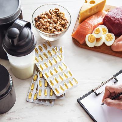 A set of food supplements such as protein powder and fish oil supplements on a table with fresh food while a person writes a diet plan.