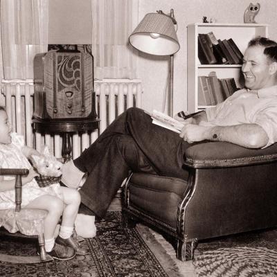 1930s Smiling father sitting in overstuffed chair watching daughter in small rocking chair both listening to wood cabinet radio.