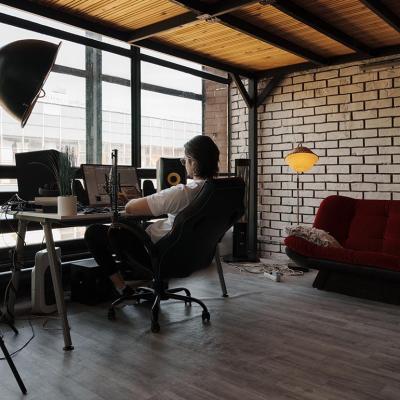 Man working on computer in a modern industrial loft office with large windows, minimalist furniture, and comfortable ambiance.