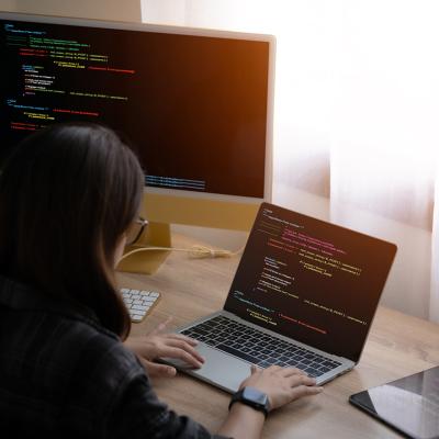 A woman working from home with a laptop and separate monitor equipment showing tech-related interface.