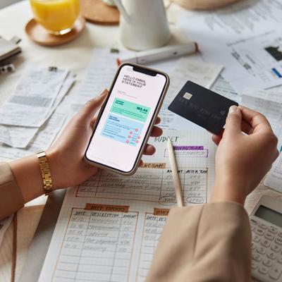 A woman organizes financial documents and is holding a credit card and a smartphone showing her statement.