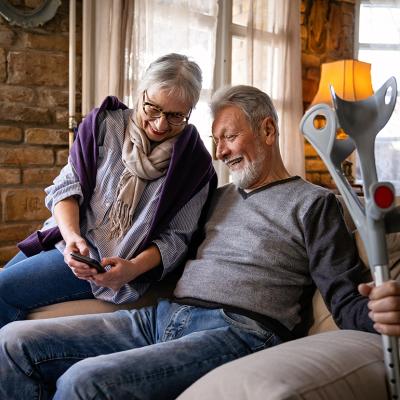 An elder couple where the wife shows her phone to her husband who is holding on to a pair of crutches while sitting on a sofa.