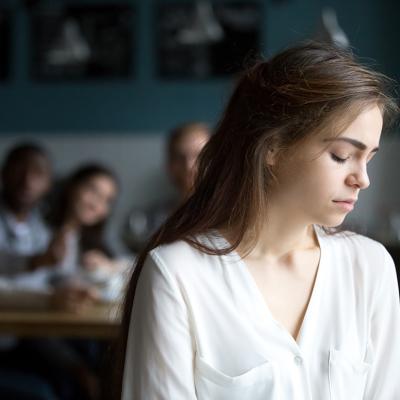A sad young woman avoiding a group peers and sitting alone in a cafe.