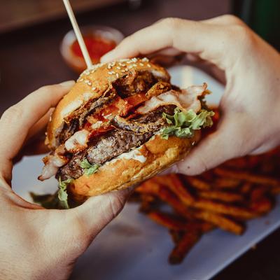 A bacon burger being held with a plate of fries in the background.