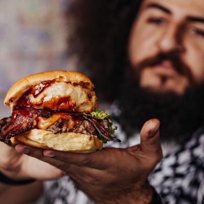 Curly hipster holding craft mouth-watering bacon and angus beef burger.