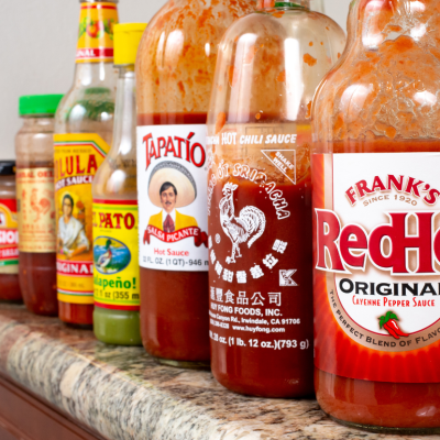 A kitchen counter with a row of popular hot sauce bottles, featuring Frank's RedHot, Huy Fong Foods sriracha, Tapatio, El Pato, and Cholula.