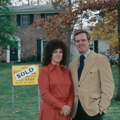 Portrait of couple standing next to For Sale sign in front of house