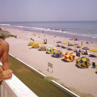 A smiling couple lean on a balcony at Thunderbird Motel, which overlooks a section of beach shaded by various colored umbrellas, Myrtle Beach, South Carolina, 1960s.