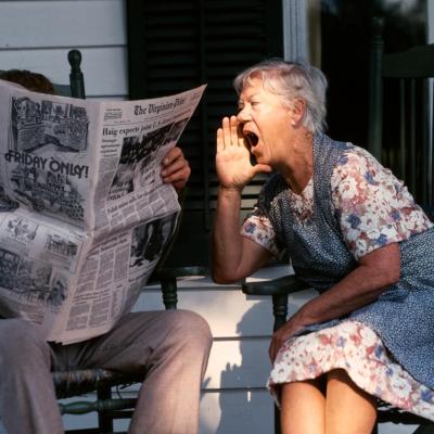 Senior couple sitting on a porch in rocking chairs. Husband is reading the paper and the wife is shouting at him. 