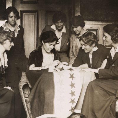 Suffragists watch as Alice Paul sews stars onto banner.