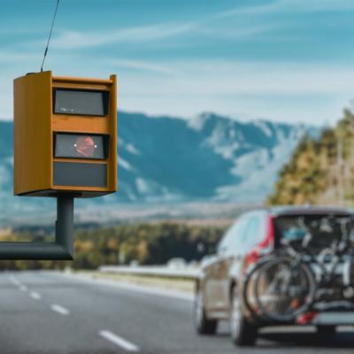 A yellow traffic speed camera prominently in the foreground, mounted on a pole along a highway.  In the background, a car with bicycles mounted on its rear.