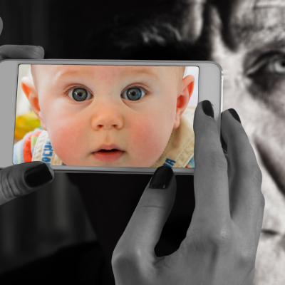 A close up of hands holding a cell phone with an image of a baby on the screen with an older man image in the background. 