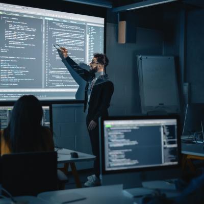 Male teacher pointing at the screen, standing in front of students in a dimly lit classroom.