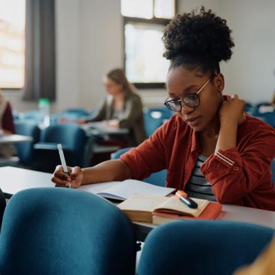 A young woman is seated at a desk. She wears glasses and a casual outfit, with a notebook, pen, and an open book on her desk. In the background, other students are seated in rows.