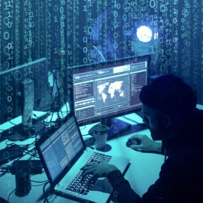 A man's silhouette in a dark room sitting in front of a laptop and another computer screen.