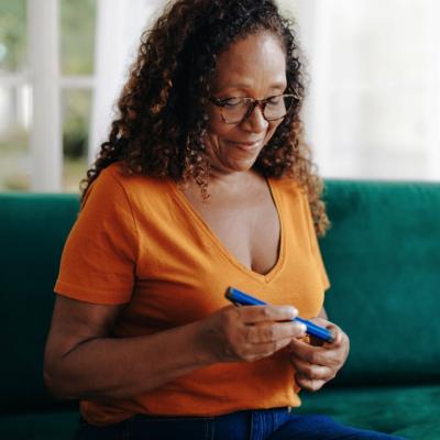 A woman sitting on a green couch, holding and looking at a blue object in her hands. The individual is wearing glasses and an orange shirt with a V-neckline. She is focused on the object they are holding.