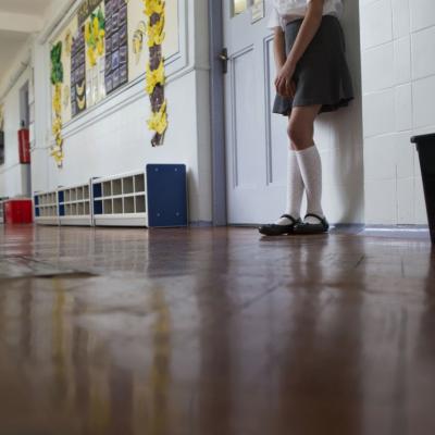 A close-up shot of a young female student's lower torso and legs. She is wearing a grey skirt, long white socks as she leans against a classroom door, standing in a school corridor.
