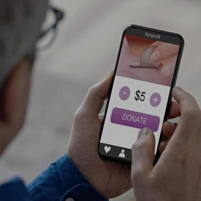 A person holding a smartphone, with a donation application on the screen displaying a coin being dropped into a slot, a selectable donation amount set to "$5" with "+" and "-" buttons and "DONATE" button.