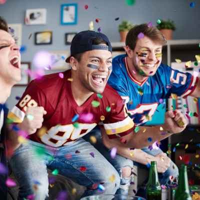 Three young men wearing football jerseys cheering while watching game at home with colorful confetti falling in the foreground. 