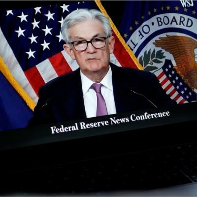 A close-up of a video or broadcast of a "Federal Reserve" news conference on a laptop screen. A man with gray hair is wearing glasses dressed in a suit with a white shirt and purple tie.