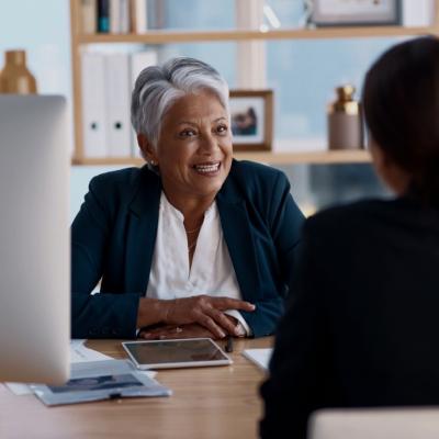 An older woman with short silver hair is looking at the woman sitting opposite her. She is wearing a dark blazer over a white blouse. She sits at a desk with various office items like a tablet, papers, and a desktop computer visible. On the right, there is another individual seen from behind.