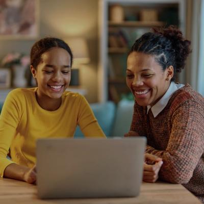 Two people sitting together at a table, both smiling warmly as they look at a laptop screen. The person on the left is a younger African American girl wearing a yellow long-sleeved shirt, looking at the laptop with a bright, happy expression. The person on the right, an African American woman, has her hair styled in a natural updo and is wearing a cozy-looking, textured sweater. 