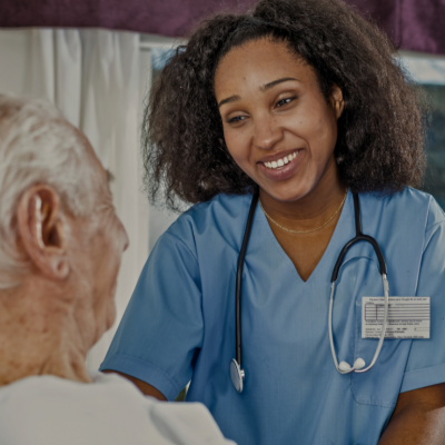 A nurse wearing blue scrubs and a stethoscope around her neck. She is smiling warmly at the patient, who is facing her and sitting up in a hospital bed.