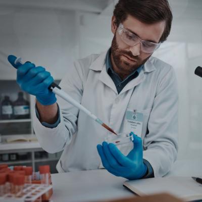 A man in white coat and protective goggles holding a petri dish and a pipette in a lab. 