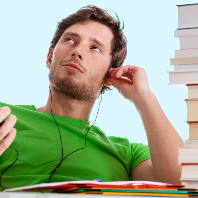 A young man sitting at the desk with a tall stack of books listining to music on his phone looking lost in his music.