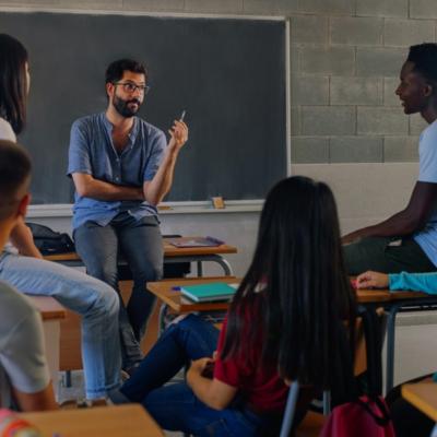 A group of students seated around a teacher in a classroom setting. The teacher casually dressed, is sitting on a desk and appears to be engaged in a discussion with the students. 