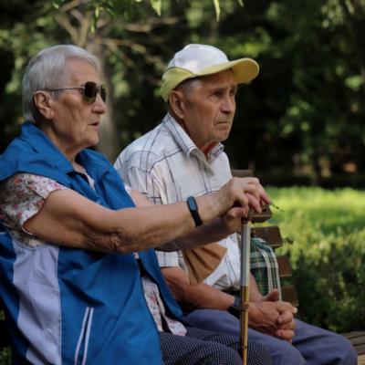 An older couple sitting on a bench in a park. An older woman wearing sunglasses, resting her hands on the cane. 