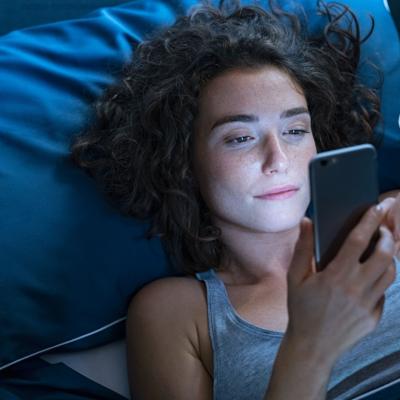 Young woman lies in bed in a dark room while looking at her phone.