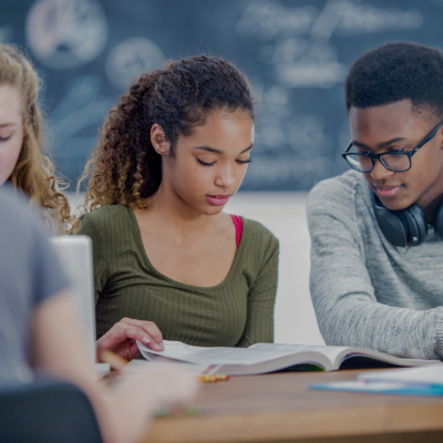 A group of diverse students sitting together in a classroom looking at the books in fornt of them. In the background, a chalkboard with various mathematical equations or diagrams visible. 