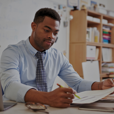 An African American male teacher is looking at a paper in his left hand. He is holding a pencil in his right hand. 