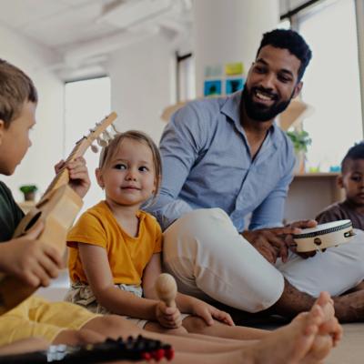 A young male teacher sitting on the floor with a tambourine in his hand surrounded by young kids.