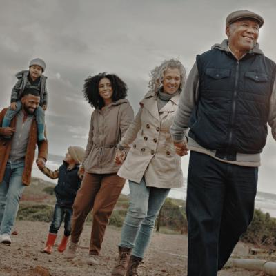 A multigenerational family taking a walk together on a beach. An older man in the front is smiling while holding hands with an older woman and behind them, a younger couple and their children. 