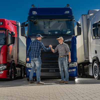 Two men in checked shirts, jeans, and baseball hats shaking hands. There are three truck cabins in the background behind them. 