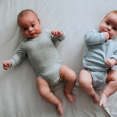 Two baby twin infants are lying down on grey sheet.