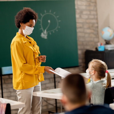 A teacher wearing a mask handing out papers to a class