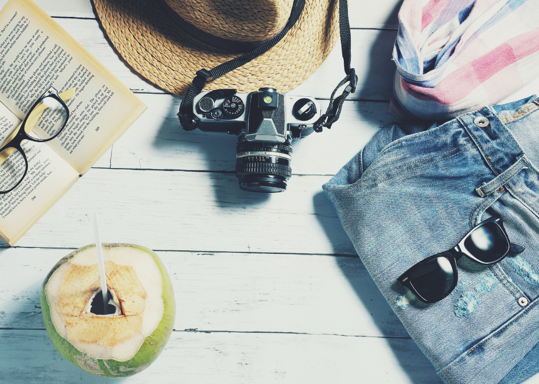 Vacation items—including a book, sunglasses, glasses, jeans, a hat, a plaid shirt, and a coconut beverage—on a white, wooden deck.