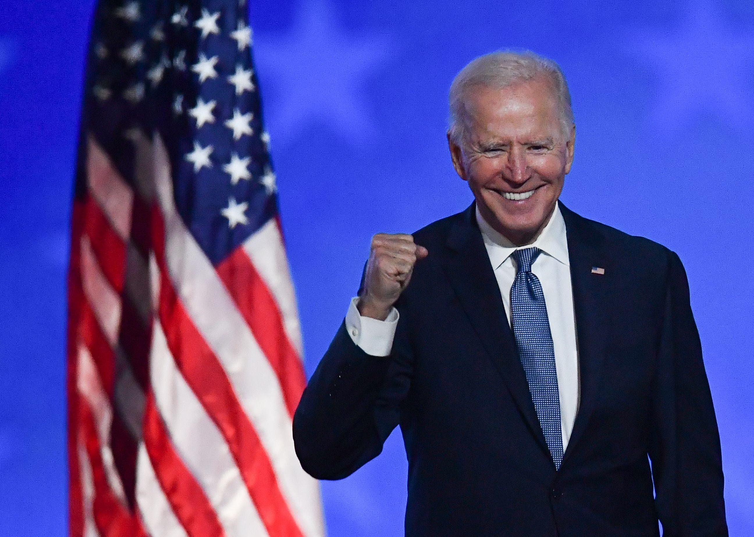 Biden standing next to an American flag smiling and cheering against a blue background.