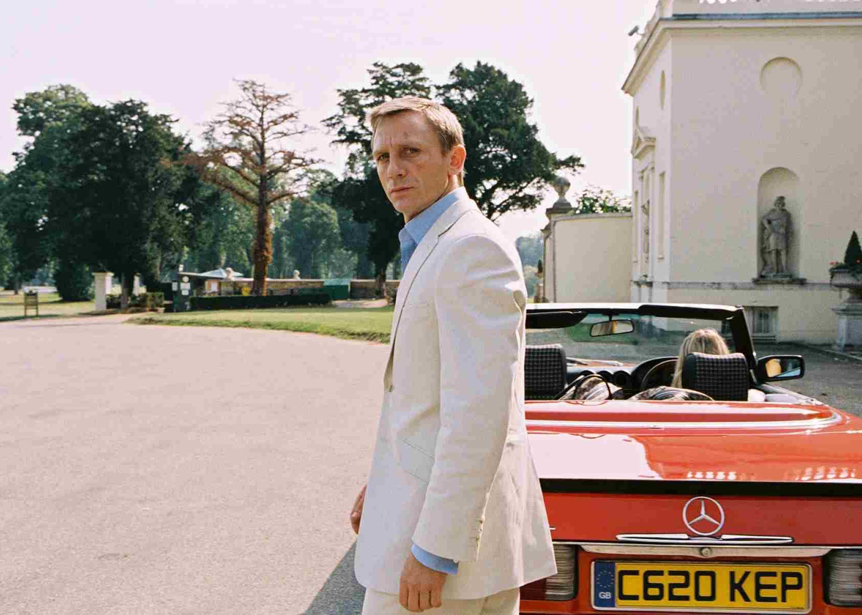 Daniel Craig in a white suit walking up to a red mercedes convertible.