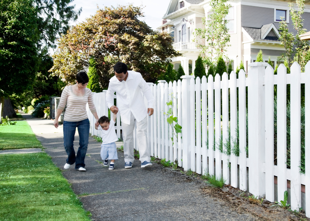 A young family walking on a sidewalk next to a white picket fence.