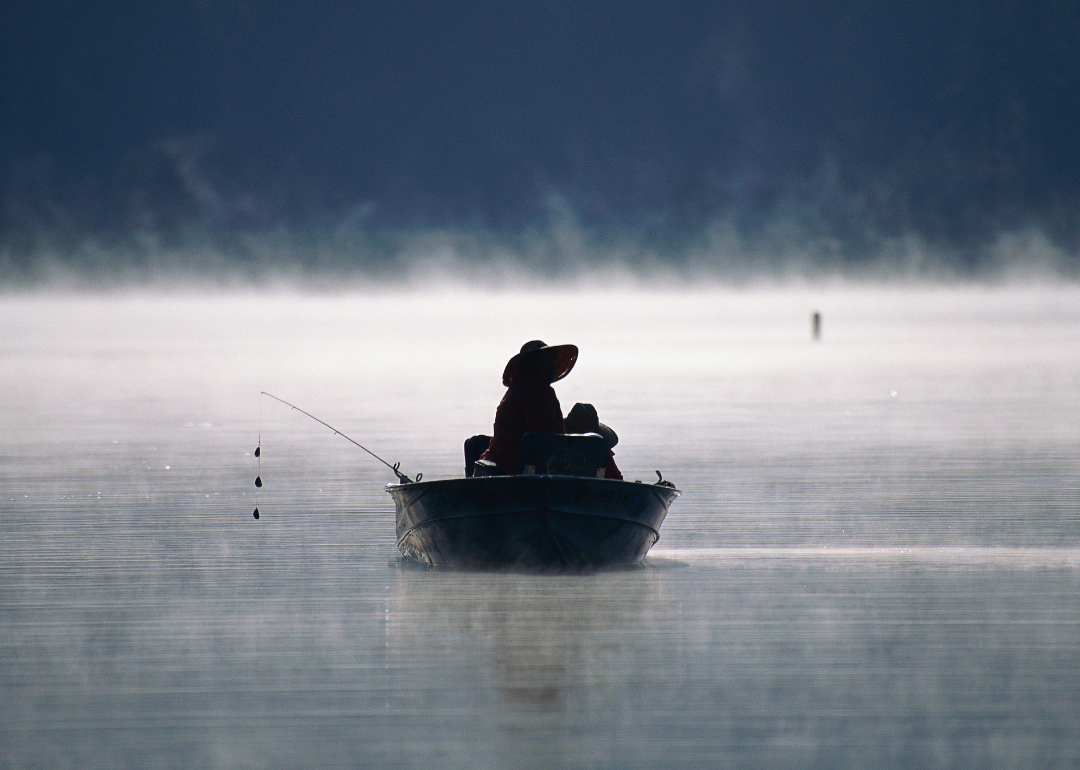 Silhouette of person sitting in a boat in a lake with a fishing pole propped on the side of the boat. 