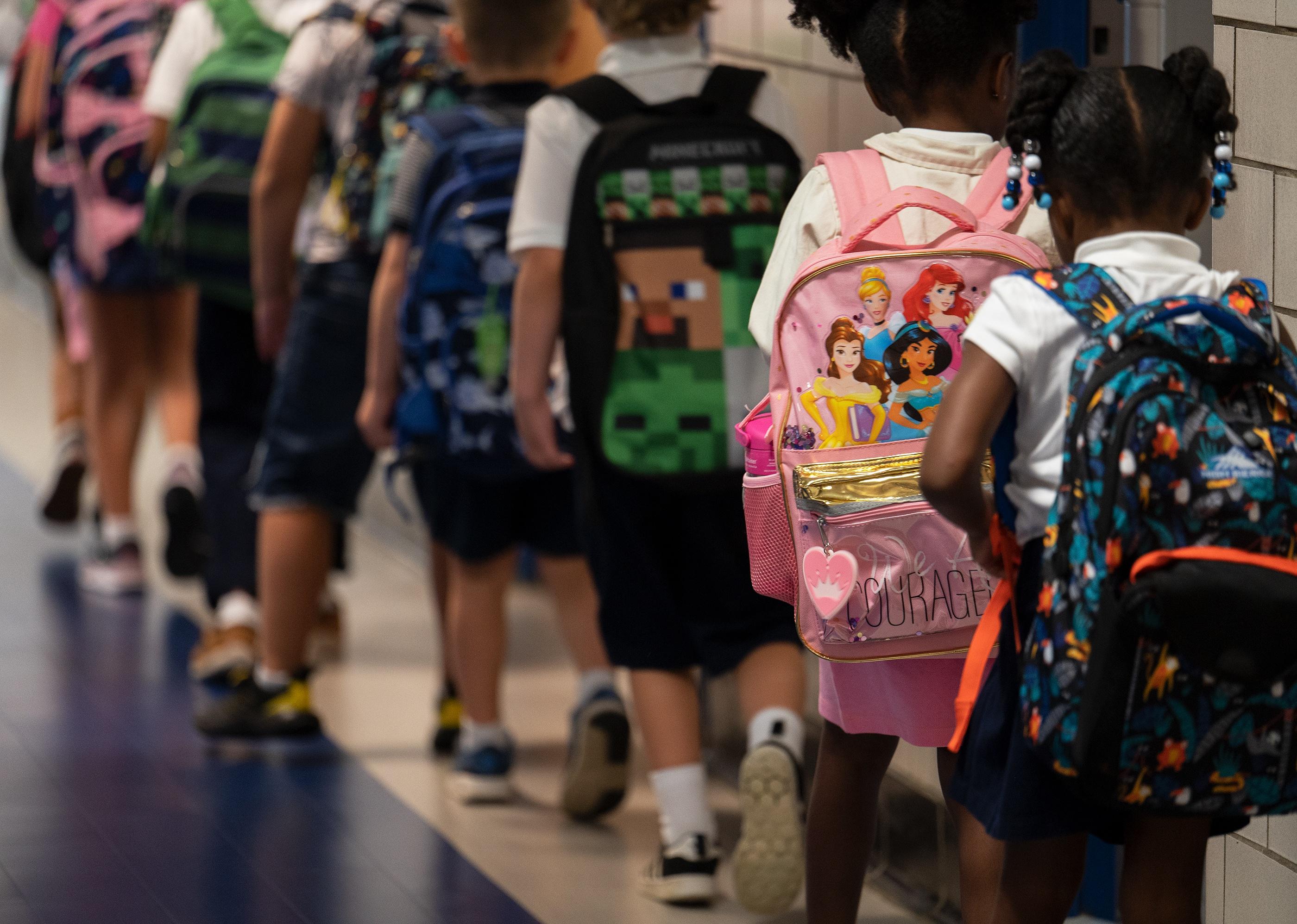 A line of kindergarten students wearing backpacks stand in a hallway at school.